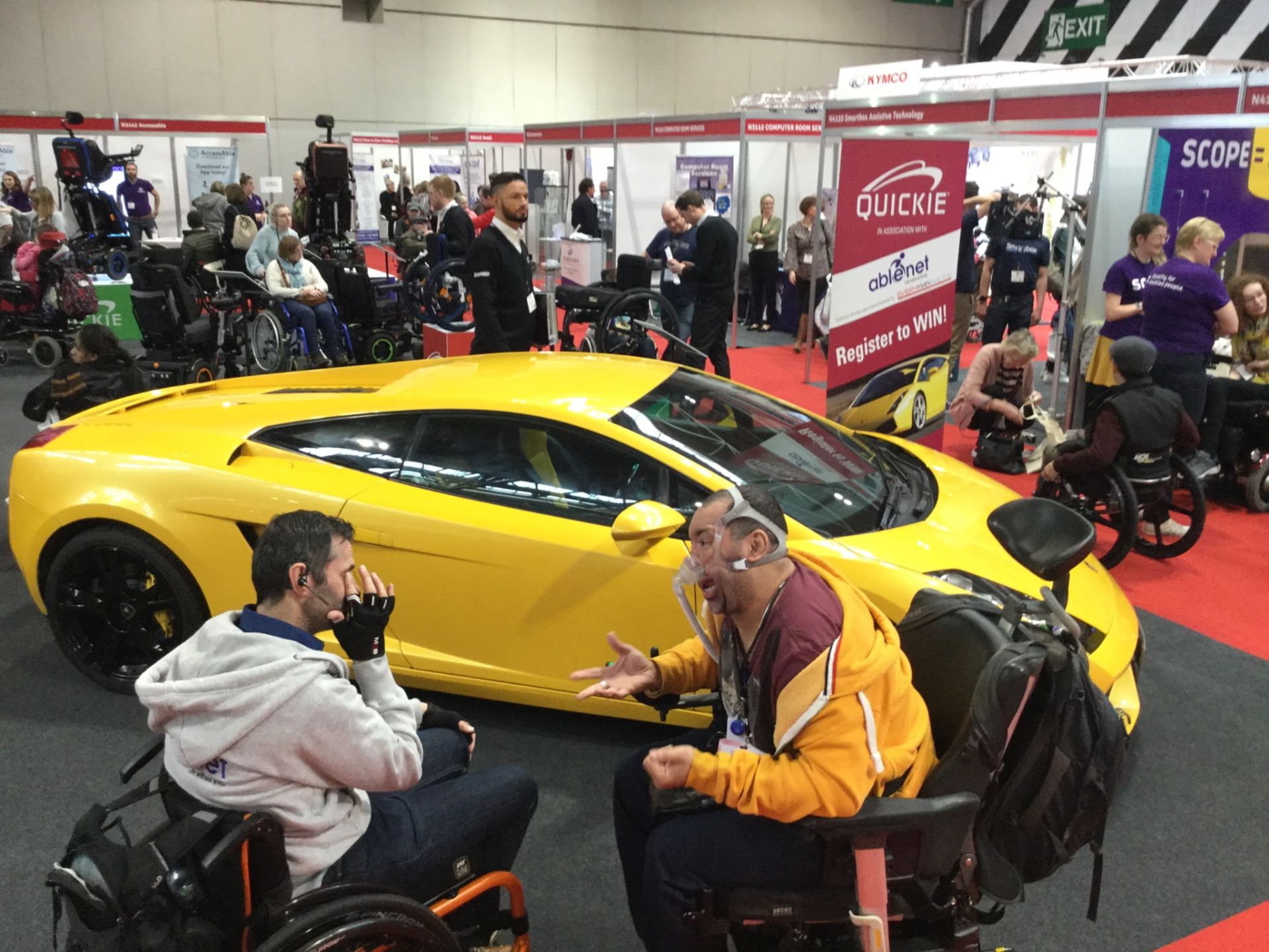 Talking with another wheelchair user at an accessibility convention, with a spectacular yellow sports car behind us.